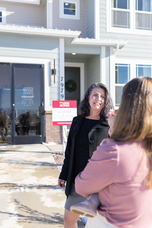 A real estate agent talking with a client outside a new build home in the snow beside a for sale sign