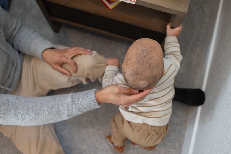 Close-up of adult hands steadying a baby learning to stand on a carpeted floor