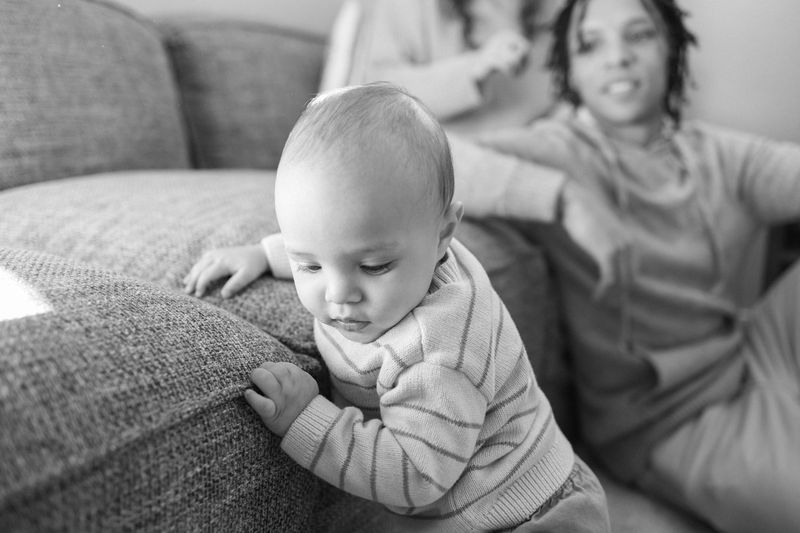 Black and white photo of a baby pulling up on a living room couch while his mother watches from behind