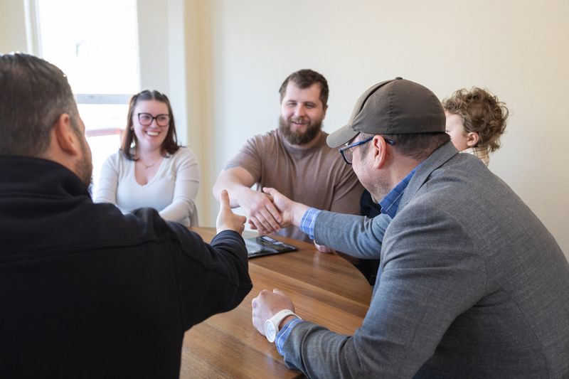Four people meeting around a wooden table, two shaking hands over an open laptop