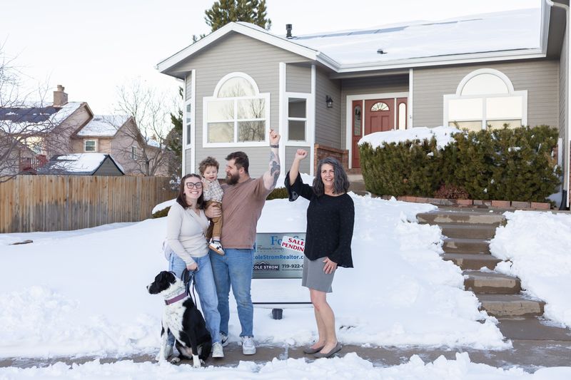 A family with their dog raising their arms in celebration in the snowy front yard of their new home beside a sale pending sign