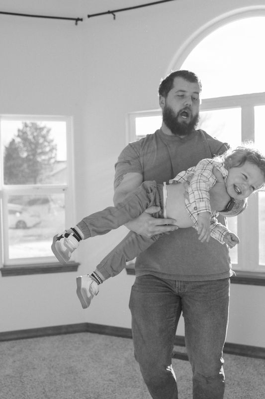 Black and white photo of a father carrying his toddler horizontally through a living room