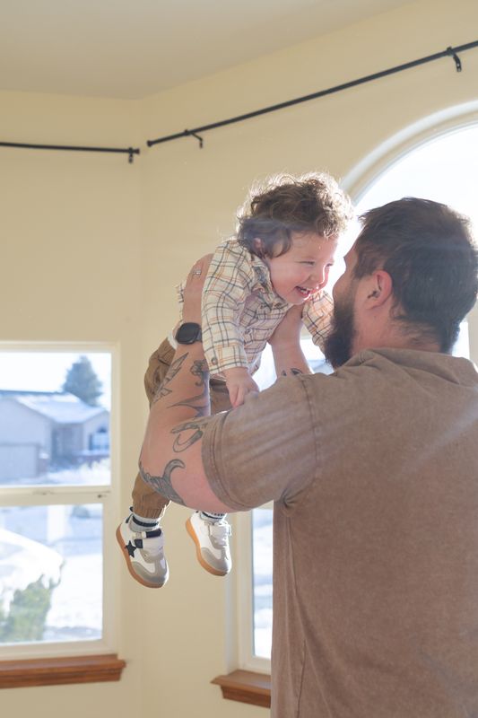 A father lifting his laughing toddler into the air in front of an arched living room window