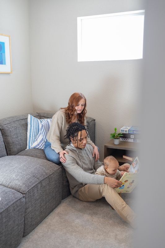 A young family of three sitting together on a grey sectional couch in a sunlit living room