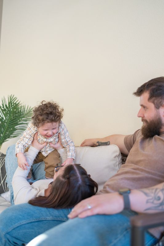 A family of three playing together on a couch, the toddler climbing over his mother