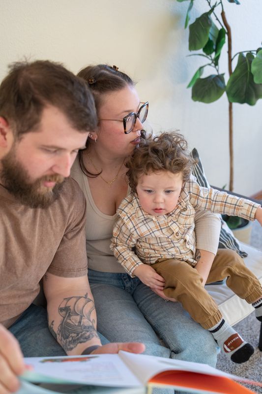 Two parents and a toddler sitting on the floor together looking at an open picture book