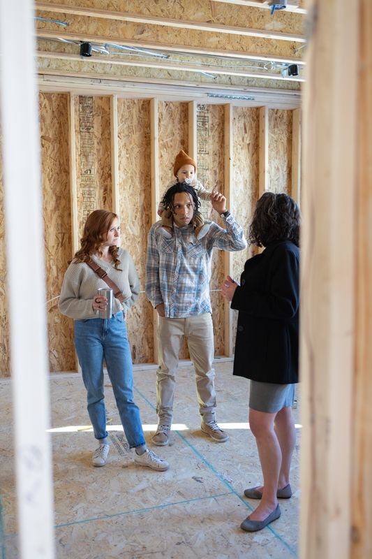 Three adults and a toddler touring a house still framed in bare studs and plywood subfloor