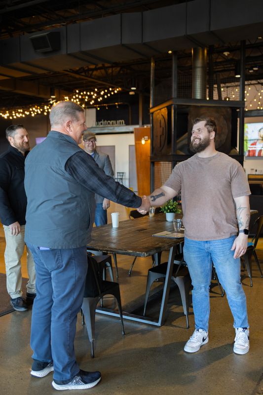 Two men shaking hands across a table in a taproom with string lights overhead