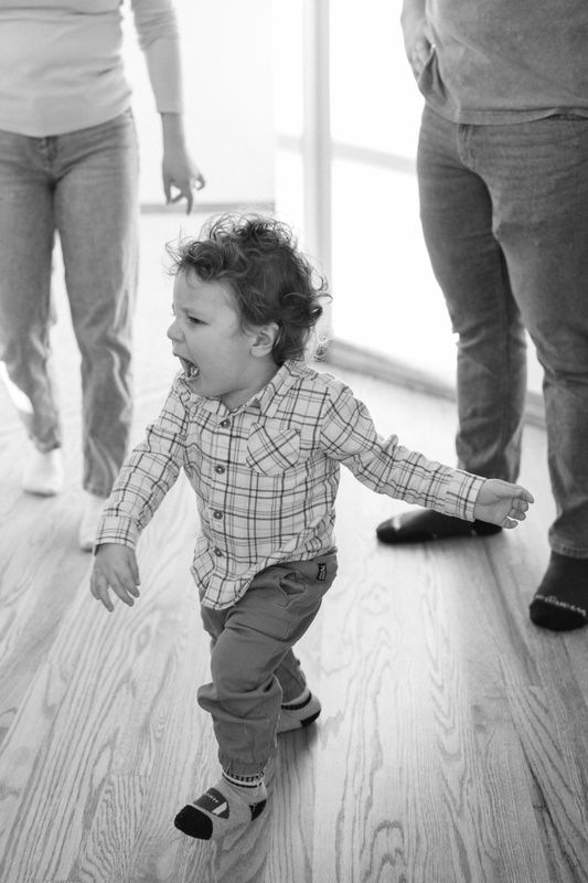 Black and white photo of a toddler walking between his parents legs across a wood floor