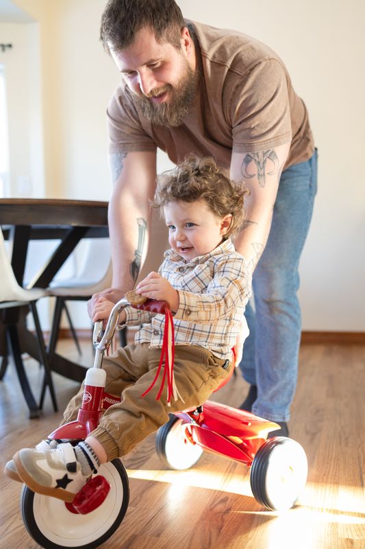 A father steadying his toddler on a red tricycle on hardwood floors indoors